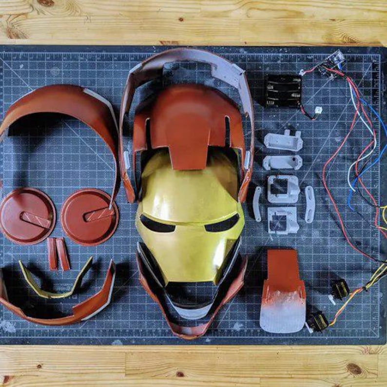 May include: A disassembled red and gold helmet kit featuring a central faceplate, various curved outer shell components, internal mounting brackets, a battery pack, and electronic wiring components laid out on a dark grid cutting mat.