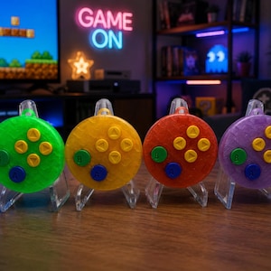 May include: Four colorful game controller-shaped ornaments. The ornaments are green, yellow, red, and purple, each with a set of yellow, green, and blue buttons. They are displayed on clear acrylic stands, with a "GAME ON" neon sign in the background.