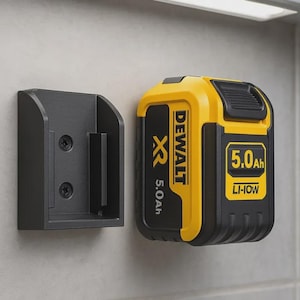 May include: A black wall-mounted battery holder next to a yellow and black DEWALT XR 5.0Ah Li-Ion battery. The battery has the text "DEWALT" and "5.0Ah" printed on it. The holder is mounted on a light gray wall.