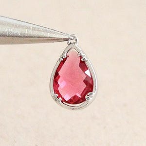 May include: A teardrop-shaped pendant featuring a faceted, ruby-coloured gemstone set in a silver-coloured metal frame. The pendant is held by tweezers against a neutral background, highlighting its intricate design and vibrant colour.