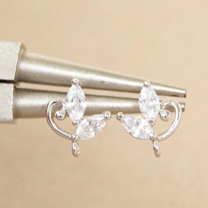 May include: A pair of silver stud earrings featuring clear, marquise-cut cubic zirconia stones. The earrings are held by a silver tool against a neutral background. The earrings have a delicate, curved design.