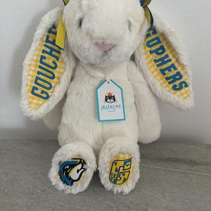 May include: A cream-colored plush rabbit with long ears featuring yellow and white gingham fabric. The ears display the blue text GOUCHER and GOPHERS. A Jellycat brand tag hangs from the neck, and the feet feature blue and yellow logos.