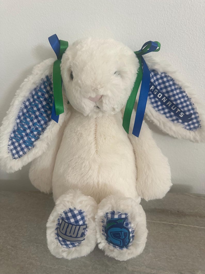 May include: A cream-colored plush rabbit with long ears featuring blue and white gingham fabric on the inner sides. The ears are tied with blue and green ribbons. The text ARGONAUTS is on one ear, and the feet feature the letters UWF and a helmet logo.