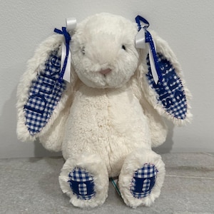 May include: A plush white rabbit toy with long ears featuring blue and white gingham fabric accents. The left ear is embroidered with the name Duke, the right with Blue Iris, and the feet feature blue gingham patches with the letter D.