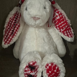 May include: A plush white rabbit toy featuring long ears lined with red and white gingham fabric. The left ear displays the text UNIVERSITY OF INDIANAPOLIS, while the right ear shows UINDY. The feet have matching gingham patches with an H and a paw print.