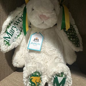 May include: A white plush rabbit toy with long ears featuring green and white checkered fabric. The ears display the text Marywood University. The rabbit has green and yellow ribbons, a Jellycat tag, and green and white checkered foot pads.