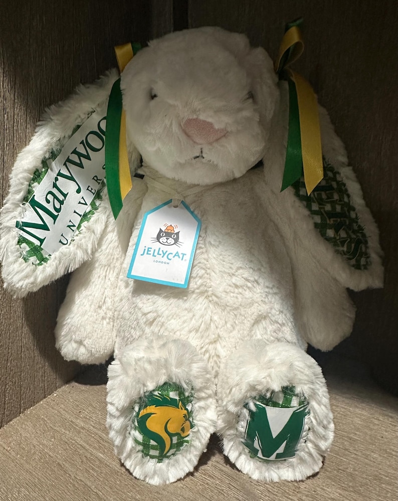 May include: A white plush rabbit toy with long ears featuring green and white checkered fabric. The ears display the text Marywood University. The rabbit has green and yellow ribbons, a Jellycat tag, and green and white checkered foot pads.