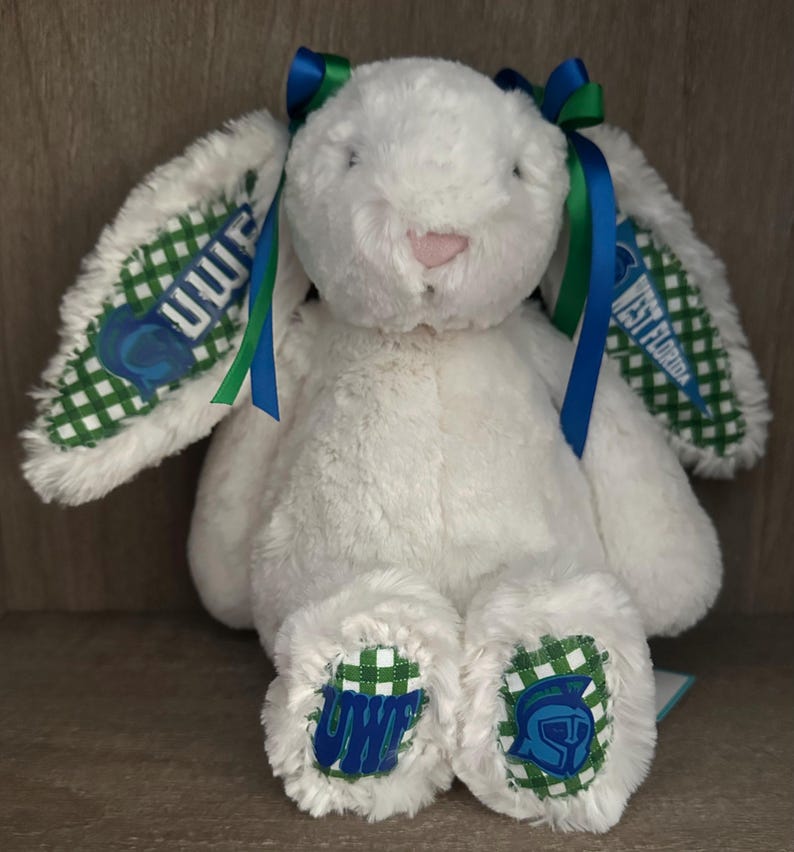 May include: A white plush rabbit toy featuring long ears with green and white gingham fabric accents. The ears and feet display the text UWF and West Florida along with a blue helmet logo. Blue and green ribbons are tied at the top of the head.