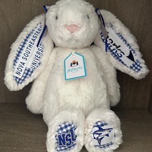 May include: A white plush rabbit toy with long ears featuring blue and white gingham fabric accents. The ears display the text Nova Southeastern University and Sharks. The feet have blue and white gingham patches with the NSU logo and a shark graphic.