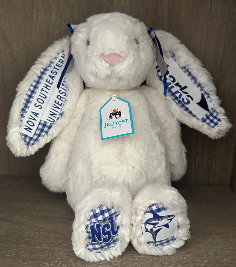 May include: A white plush rabbit toy with long ears featuring blue and white gingham fabric accents. The ears display the text Nova Southeastern University and Sharks. The feet have blue and white gingham patches with the NSU logo and a shark graphic.