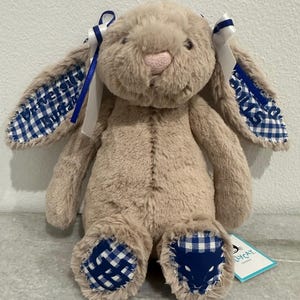 May include: A beige plush rabbit with long ears featuring blue and white gingham fabric lining. The ears are embroidered with the text University Buffalo. The rabbit has blue and white ribbons on its ears and blue and white gingham foot pads.