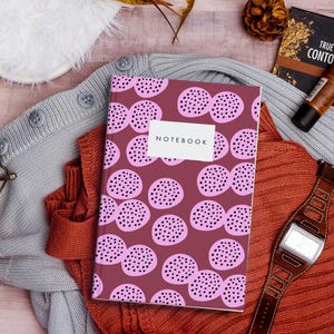 Puede incluir: Un cuaderno con una cubierta burdeos con un patrón de puntos rosas y la palabra «NOTEBOOK» en una etiqueta blanca. El cuaderno descansa sobre un suéter color óxido, con un suéter gris y gafas cerca. También se ve un reloj marrón y maquillaje.