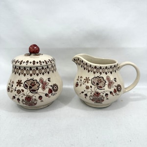 May include: Cream-colored ceramic sugar bowl with lid and creamer set. The set features a floral pattern in shades of brown and red. The sugar bowl has a round shape with a lid and a red knob. The creamer has a handle and a spout.