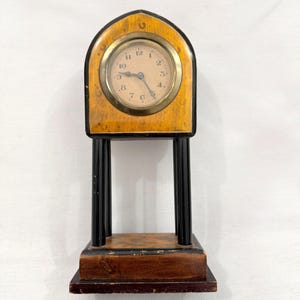May include: A mantel clock featuring a light brown wood-grained arched top with a brass-rimmed circular face, black numerals, and two hands. The clock is supported by four slender black pillars resting on a rectangular dark wood base.