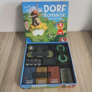 May include: A Dorfromantik board game box with a windmill illustration and a 2023 Spiel des Jahres award seal. Below, an open box contains black 3D-printed organizers with labels reading Hexes, Hex Holders, Markers, Flags, and Boxes for game components.