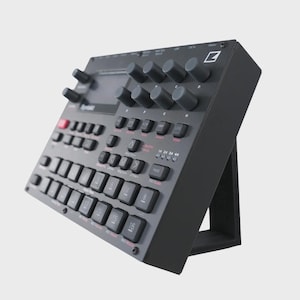 May include: A dark grey electronic music production unit featuring multiple knobs, buttons, and a small display screen. The device is propped up on a stand, displaying its interface. The buttons are labelled with various functions, and the design is sleek and contemporary.