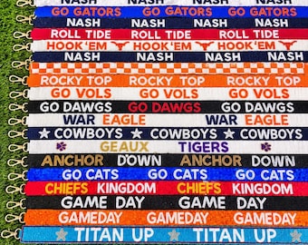 May include: A collection of fifteen beaded bag straps featuring various sports-themed text and slogans including NASH, GO GATORS, ROLL TIDE, HOOK 'EM, ROCKY TOP, GO VOLS, GO DAWGS, WAR EAGLE, COWBOYS, GEAUX TIGERS, ANCHOR DOWN, GO CATS, CHIEFS KINGDOM, GAME DAY, and TITAN UP.