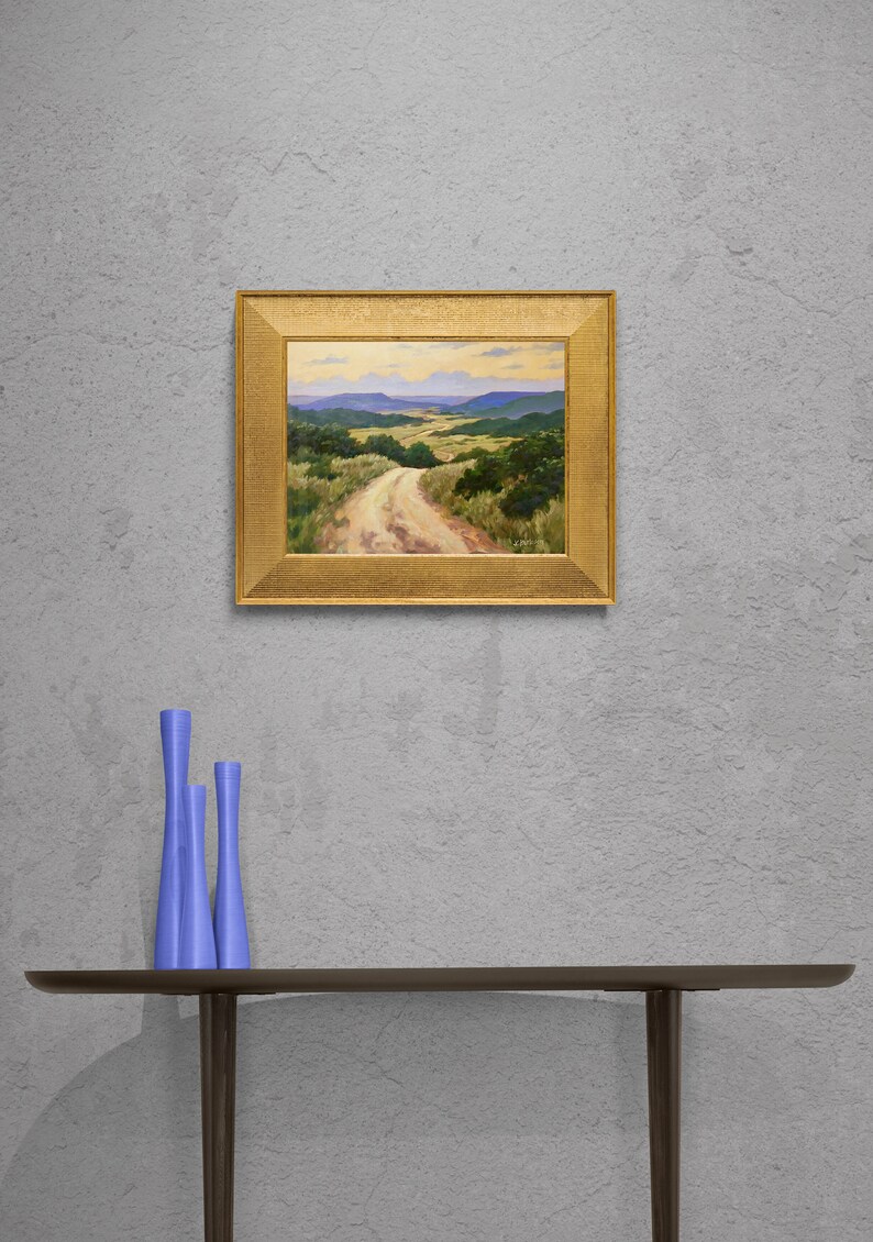 May include: A framed landscape painting in a gold ornate frame. The painting shows a dirt track winding through a valley with green hills and blue mountains. Three blue decorative vases are on a dark brown shelf.