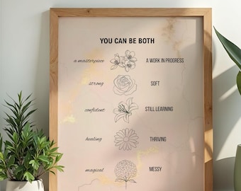 May include: A framed print with the text "YOU CAN BE BOTH" and various descriptive words paired with floral illustrations. The frame is light brown, and the background is a soft pink with gold accents. The print is leaning against a white wall.