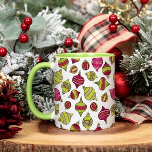 May include: A white ceramic mug with a lime green interior and handle, featuring a repeating pattern of illustrated Christmas ornaments in shades of magenta, lime green, and yellow with black outlines against a plain white background.