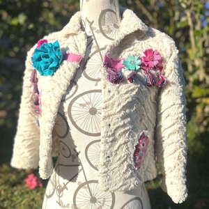 May include: A cream-colored textured jacket featuring a large teal fabric flower on the left lapel, pink ribbon trim, and decorative accents including a light blue flower, a pink flower, and small hanging tassels on the right side of the collar.