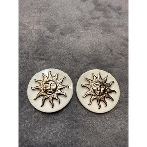 May include: A pair of round earrings featuring a sun design. The earrings have a white background with a metallic gold sun face and rays. The earrings are set against a gray background.