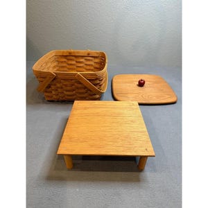 May include: A light brown woven basket with a handle, a wooden square lid with a small red apple, and a small wooden table with four legs. The items are made of wood and are arranged on a gray surface.