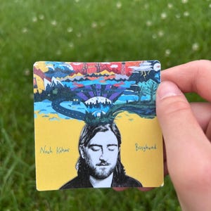 May include: A square, yellow card featuring a portrait of a man with long hair and a beard. The card has the text "Noah Kahan" and "Busyhead". The top of the card has a colorful illustration of a landscape with mountains, a road, and a city.
