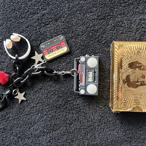 May include: A selection of keyrings and a gold-coloured box. The keyrings feature a white headset, a red and black cassette tape, a grey boombox, a red pom-pom, and star charms. The gold box has a face design and the number 100.
