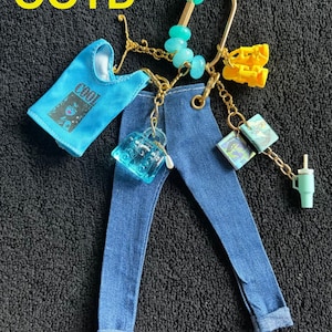 Puede incluir: Llavero dorado con mini vaqueros, una camiseta sin mangas azul con el texto "COOL" y varios accesorios. Estos incluyen un pequeño bolso, libros, un objeto amarillo, cuentas turquesas y una pequeña taza verde.
