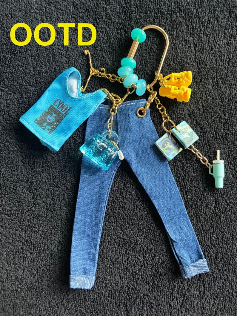 Op de afbeelding: Gouden sleutelhanger met miniatuur spijkerbroek, een blauwe tanktop met de tekst "COOL" en diverse accessoires. Deze omvatten een kleine handtas, boeken, een geel item, turkooizen kralen en een klein groen bekertje.