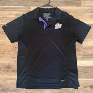 May include: A black short-sleeved polo shirt featuring a Reebok logo on the sleeve and a PlayDry label at the hem. The collar placket is lined with purple fabric, and the chest displays a colorful graphic of a hockey player with the text Sun Devils.