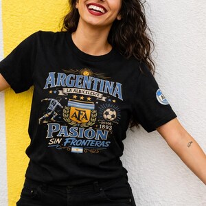 以下が含まれることがあります： 青と金の「ARGENTINA」の文字と「PASION SIN FRONTERAS」の文字が描かれたグラフィックデザインの黒いTシャツ。シャツにはサッカーボールとAFAのロゴも含まれています。