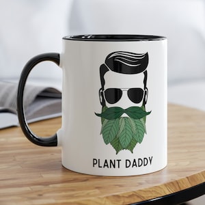 Puede incluir: Taza de cerámica blanca con borde y asa negros. La taza presenta una ilustración en blanco y negro de una persona con gafas de sol y una barba verde frondosa. El texto "PLANT DADDY" está impreso debajo de la ilustración.