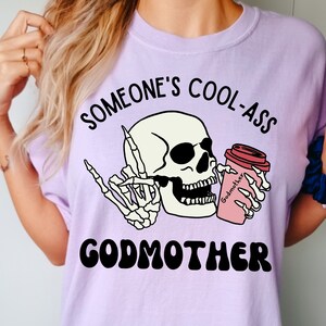 May include: A purple t-shirt with the text "Someone's Cool-Ass Godmother" in black lettering. A cartoon skull with sunglasses is holding a pink coffee cup with the text "Godmother" on it.
