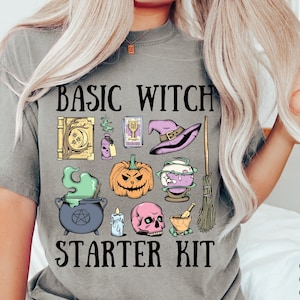 May include: A grey t-shirt featuring the text "Basic Witch Starter Kit" and a graphic of assorted witchy items, such as a cauldron, a broom, a skull, a pumpkin, and a book. The text "Comfort Colours: Grey" is also visible on the shirt.