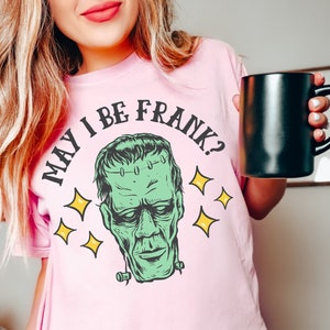 May include: A light pink t-shirt featuring a green Frankenstein's monster graphic with the text "MAY I BE FRANK?" in a black, circular font. The design includes yellow star accents. The shirt is a Comfort Colors brand.