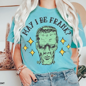 May include: A light blue t-shirt with the text "MAY I BE FRANK?" in black, featuring a green Frankenstein's monster graphic with yellow star accents. The shirt is a comfort color in chalky mint.