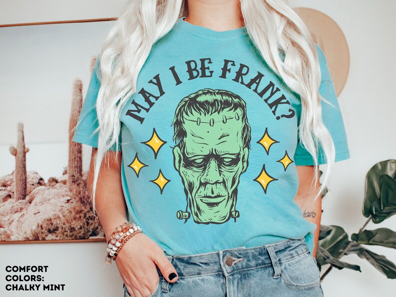 May include: A light blue t-shirt with the text "MAY I BE FRANK?" in black, featuring a green Frankenstein's monster graphic with yellow star accents. The shirt is a comfort color in chalky mint.