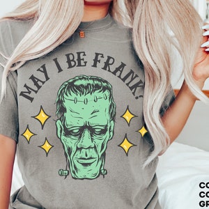 May include: A grey t-shirt featuring a green Frankenstein's monster graphic with the text "MAY I BE FRANK?" and yellow star accents. The shirt is part of the Comfort Colors collection and is grey.