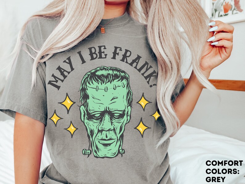 May include: A grey t-shirt featuring a green Frankenstein's monster graphic with the text "MAY I BE FRANK?" and yellow star accents. The shirt is part of the Comfort Colors collection and is grey.