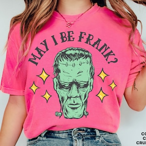May include: A bright pink t-shirt with the phrase "MAY I BE FRANK?" in black arched text above a green Frankenstein's monster graphic. Yellow starbursts surround the graphic. The shirt is a Comfort Colors brand in the color Crunchberry.