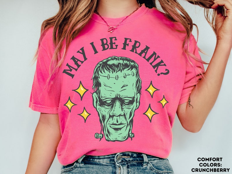 May include: A bright pink t-shirt with the phrase "MAY I BE FRANK?" in black arched text above a green Frankenstein's monster graphic. Yellow starbursts surround the graphic. The shirt is a Comfort Colors brand in the color Crunchberry.