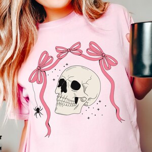 K&ouml;nnte beinhalten: Hellrosa T-Shirt mit einem Totenkopf-Design, rosa Schleifen und einer Spinne. Der Sch&auml;del ist cremefarben mit schwarzen Augenh&ouml;hlen. Der Text "COMFORT COLORS: BLOSSOM" steht unten.