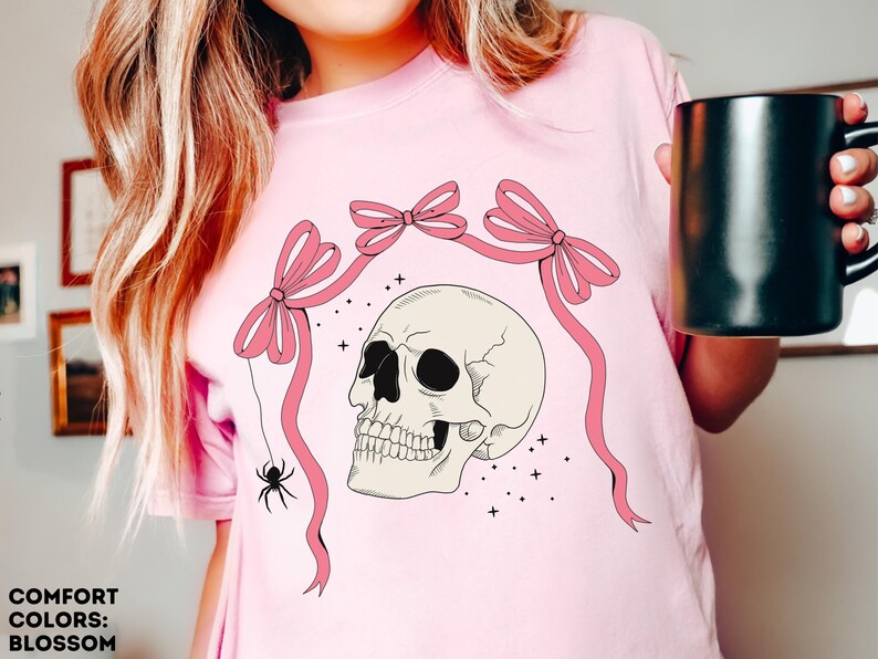 K&ouml;nnte beinhalten: Hellrosa T-Shirt mit einem Totenkopf-Design, rosa Schleifen und einer Spinne. Der Sch&auml;del ist cremefarben mit schwarzen Augenh&ouml;hlen. Der Text "COMFORT COLORS: BLOSSOM" steht unten.