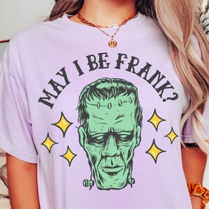 May include: Orchid-colored t-shirt featuring a green Frankenstein's monster graphic with the phrase "MAY I BE FRANK?" in black. The design includes yellow star accents. The shirt is a comfort color.