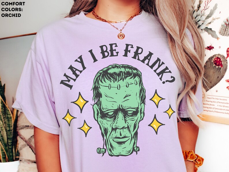 May include: Orchid-colored t-shirt featuring a green Frankenstein's monster graphic with the phrase "MAY I BE FRANK?" in black. The design includes yellow star accents. The shirt is a comfort color.