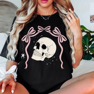 K&ouml;nnte beinhalten: Schwarzes T-Shirt mit einem Totenkopf-Design, rosa Schleifen und einer Spinne. Der Text "COMFORT COLORS: BLACK" ist sichtbar. Das Design ist vor einem Sternenhintergrund platziert. Ein Gothic-Themen-Kleidungsst&uuml;ck.