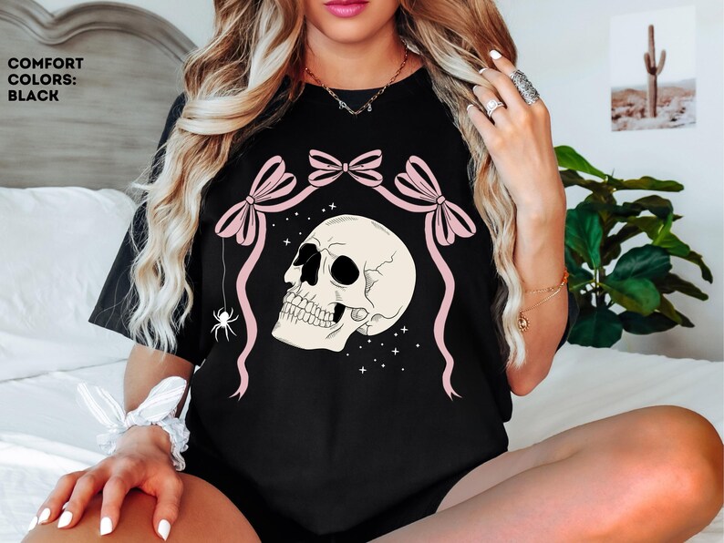 K&ouml;nnte beinhalten: Schwarzes T-Shirt mit einem Totenkopf-Design, rosa Schleifen und einer Spinne. Der Text "COMFORT COLORS: BLACK" ist sichtbar. Das Design ist vor einem Sternenhintergrund platziert. Ein Gothic-Themen-Kleidungsst&uuml;ck.