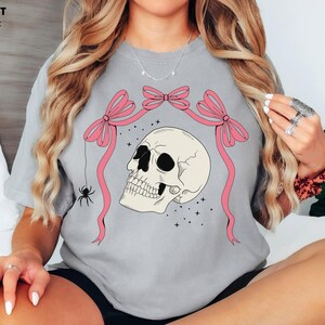 K&ouml;nnte beinhalten: Graues T-Shirt mit einem Totenkopf-Motiv. Der Totenkopf ist cremefarben mit schwarzen Augenh&ouml;hlen und einem detaillierten Kiefer. Rosa Schleifen und B&auml;nder umrahmen den Totenkopf, mit einer Spinne, die an einem Band h&auml;ngt. Der Text "COMFORT COLORS: GREY" steht oben links.